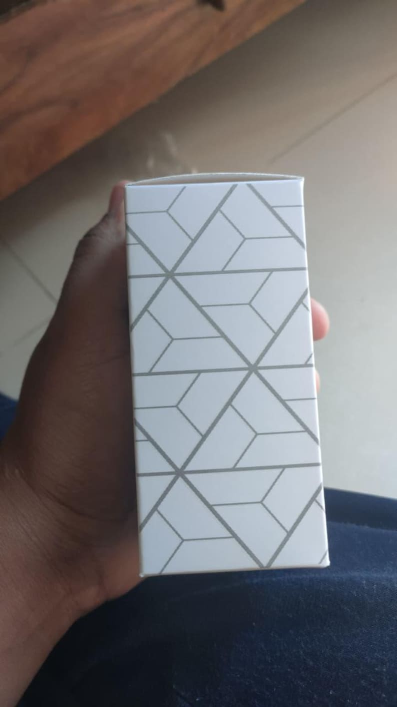 Puede incluir: Una caja rectangular blanca con un patr&oacute;n geom&eacute;trico gris. La caja est&aacute; sostenida por una persona. El fondo est&aacute; desenfocado.