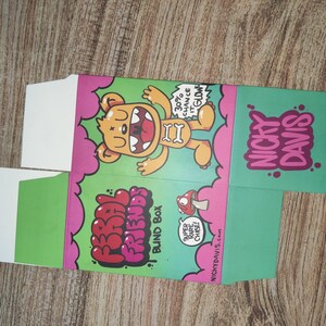 Puede incluir: Una caja de cart&oacute;n plegada con una ilustraci&oacute;n de un oso de dibujos animados. La caja es verde, rosa y blanca, con el texto "FRIENDS BLIND BOX" y "NICKY DAVIS". El oso es amarillo con un hueso en la boca. La caja tambi&eacute;n tiene el texto "30% chance it glows".