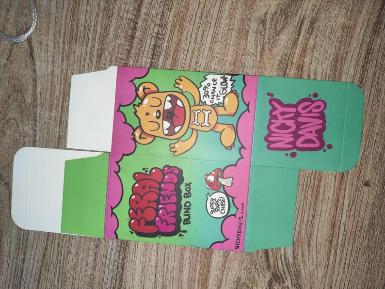 Puede incluir: Una caja de cart&oacute;n plegada con una ilustraci&oacute;n de un oso de dibujos animados. La caja es verde, rosa y blanca, con el texto "FRIENDS BLIND BOX" y "NICKY DAVIS". El oso es amarillo con un hueso en la boca. La caja tambi&eacute;n tiene el texto "30% chance it glows".