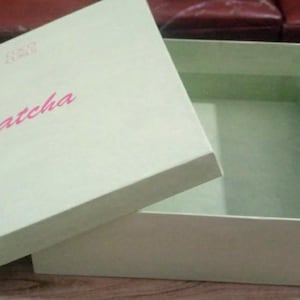 Puede incluir: Caja verde claro abierta, revelando el interior. La tapa tiene la palabra "matcha" en rosa. La caja está sobre una superficie de madera. Ideal para embalaje o almacenamiento.