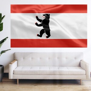 May include: A large wall hanging featuring the flag of Berlin, Germany. The flag has a red top and bottom stripe with a white center stripe. A black bear is centered on the white stripe. The flag is displayed above a cream-colored sofa.