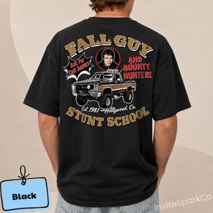 May include: A black t-shirt featuring a graphic on the back with a vintage truck, a portrait of a man, and the text "FALL GUY AND BOUNTY HUNTERS STUNT SCHOOL", "Ask For Colt Seavers", and "Est. 1981 Hollywood, Ca." printed in tan, red, and white.