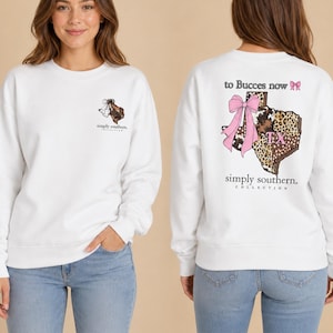 May include: A white long-sleeve sweatshirt featuring a small graphic on the front left chest and a large graphic on the back. The back design shows a map of Texas with animal print patterns, a large pink bow, and the text to Bucces now TX.
