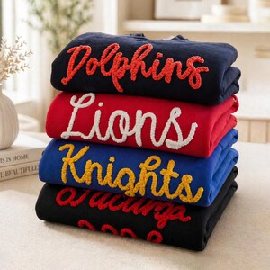 May include: A stack of four folded sweatshirts in navy, red, blue, and black. Each sweatshirt features textured, raised lettering in a contrasting color. The top sweatshirt reads Dolphins in red, followed by Lions in white, Knights in yellow, and a black one.