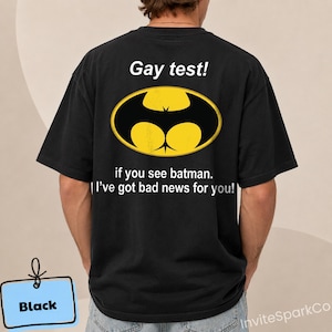 May include: A black short-sleeved t-shirt featuring a yellow and black oval graphic resembling a bat symbol. The text reads: Gay test! if you see batman. I've got bad news for you! The shirt is displayed from the back, showing the printed design.