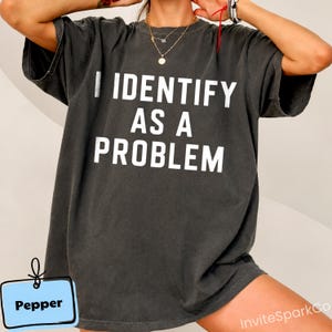 May include: A charcoal grey short-sleeve t-shirt featuring the white text I IDENTIFY AS A PROBLEM printed in a bold, sans-serif font across the chest. The garment has a relaxed fit with a crew neckline and a slightly faded, vintage-style finish.