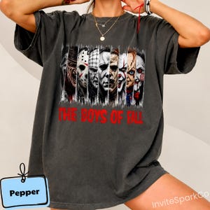 May include: A charcoal gray short-sleeve t-shirt featuring a graphic print of various horror movie characters arranged in vertical panels. Below the characters, the red text reads THE BOYS OF FALL. The shirt has a relaxed, loose-fitting silhouette.