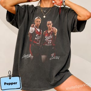 May include: A charcoal gray short-sleeve t-shirt featuring a graphic print of two basketball players in red jerseys with the text Indy 8 and Indy 22. The design includes two white signatures at the bottom and a small blue tag labeled Pepper.