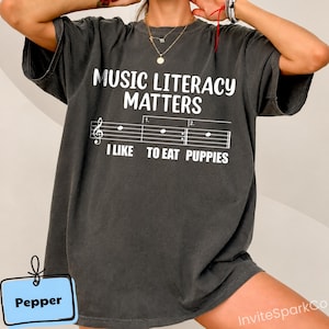 May include: A charcoal grey short-sleeve t-shirt featuring the white text MUSIC LITERACY MATTERS, a musical staff with notes, and the text I LIKE TO EAT PUPPIES. A small light blue rectangular tag in the corner displays the word Pepper.
