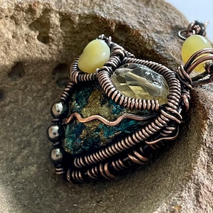 May include: A copper wire wrapped pendant featuring a green and gold stone, a clear quartz crystal, and two yellow-green stones. The pendant is resting on a rough, gray stone surface.