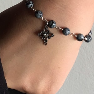 May include: A silver chain bracelet with gray and black speckled beads and a silver charm in the shape of a cross.