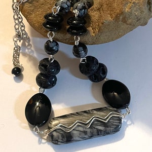 May include: A silver chain necklace with black and grey gemstone beads. The necklace features a long, rectangular grey stone with silver wire wrapped around it.