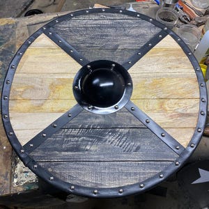 Op de afbeelding: Een rond houten schild met een zwarte metalen rand en een centrale zwarte schildknop. Het schild heeft afwisselend lichte en donkere houten panelen, zwarte leren riemen en zilveren klinknagels. Een decoratief item.