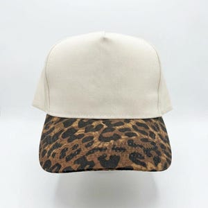May include: A beige baseball cap with a leopard print brim. The cap has a structured, flat brim and a rounded crown. The leopard print features brown and black spots on a tan background. The cap is set against a white background.