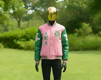 Op de afbeelding: Een roze en groene varsity jas met de letters "AKA" en een embleem op de voorkant. De jas heeft groene mouwen en biezen, met de letters "AKA" in het wit. De jas wordt getoond op een mannequin in een grasveld.