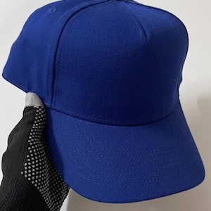 Può includere: Un cappellino da baseball blu reale con visiera curva. Il cappello è realizzato in un tessuto strutturato e presenta un bottone sulla parte superiore. Il cappellino è tenuto da una mano che indossa un guanto nero con pois bianchi.