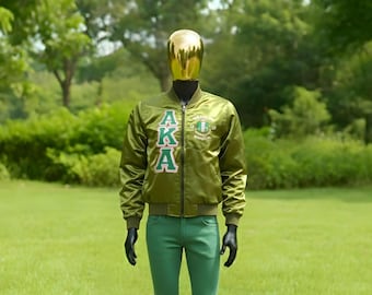 Op de afbeelding: Een groene satijnen bomberjack met de letters "AKA" in wit en groen, gecombineerd met een groene broek. De jas heeft een ritssluiting en geribde manchetten. De etalagepop staat tegen een achtergrond van groen gras en bomen.