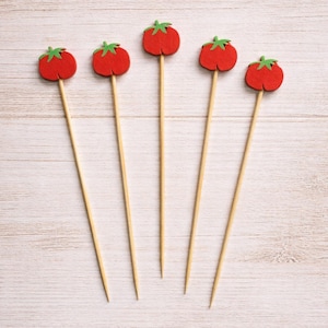 May include: Five decorative food picks featuring small, red, felt-like tomato shapes with green stems attached to the tops of thin, pointed wooden skewers. The skewers are arranged in a fan-like pattern, showcasing the uniform design of each pick.