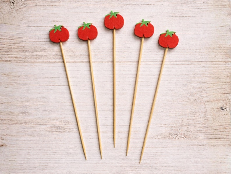 May include: Five decorative food picks featuring small, red, felt-like tomato shapes with green stems attached to the tops of thin, pointed wooden skewers. The skewers are arranged in a fan-like pattern, showcasing the uniform design of each pick.