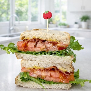 May include: A double-decker BLT sandwich featuring layers of toasted bread, crisp green lettuce, sliced red tomatoes, and cooked bacon. A small wooden toothpick with a red tomato-shaped topper is inserted into the top slice of bread.