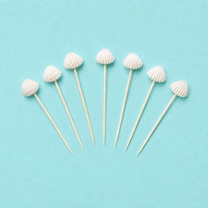 May include: Seven wooden cocktail sticks, each topped with a small, white, ridged seashell. The sticks are arranged in a fan shape, with the pointed ends directed downwards, showcasing the natural light wood texture and the detailed texture of the shells.