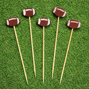 May include: Five brown felt American football toppers attached to thin wooden skewers. Each football features white stitched detailing across the center. The toppers are arranged in a row, showcasing the brown texture and white accents clearly.