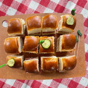 May include: A dozen golden-brown slider sandwiches arranged on a rectangular brown cutting board. Each sandwich is topped with a small green decorative pick featuring a slice of pickle. The sliders are positioned in a grid pattern on the board.