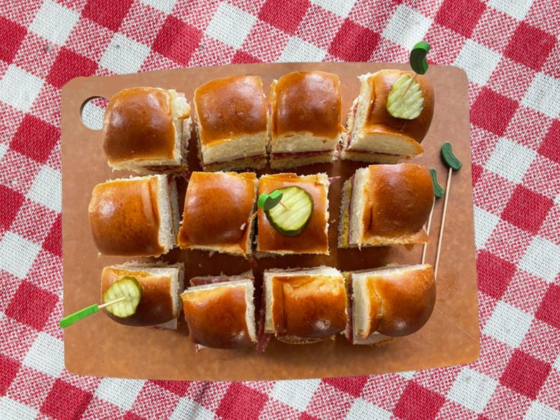 May include: A dozen golden-brown slider sandwiches arranged on a rectangular brown cutting board. Each sandwich is topped with a small green decorative pick featuring a slice of pickle. The sliders are positioned in a grid pattern on the board.