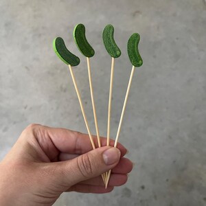 May include: Four wooden skewers, each topped with a small, curved, textured green pickle-shaped ornament. The skewers are thin, light-colored, and pointed at the bottom, featuring a flat, decorative green pickle topper at the top end of each stick.