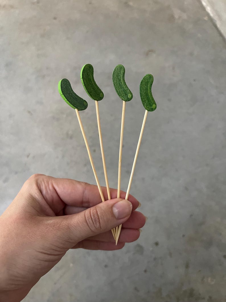 May include: Four wooden skewers, each topped with a small, curved, textured green pickle-shaped ornament. The skewers are thin, light-colored, and pointed at the bottom, featuring a flat, decorative green pickle topper at the top end of each stick.