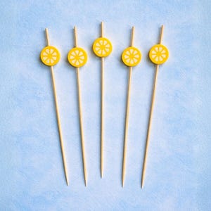 May include: Five wooden cocktail sticks, each topped with a yellow lemon slice. The lemon slices feature a white starburst pattern. The sticks are arranged on a light blue textured surface.