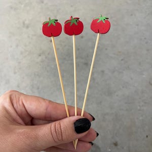 May include: Three wooden food picks, each topped with a flat, red tomato-shaped cutout featuring a small green stem detail. The picks are made of light-colored, thin, pointed wooden sticks, designed for serving appetizers or garnishing food items.