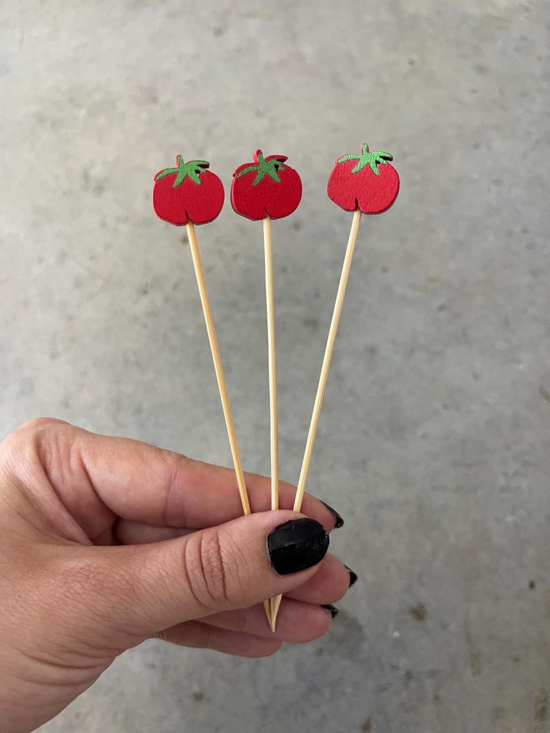 May include: Three wooden food picks, each topped with a flat, red tomato-shaped cutout featuring a small green stem detail. The picks are made of light-colored, thin, pointed wooden sticks, designed for serving appetizers or garnishing food items.