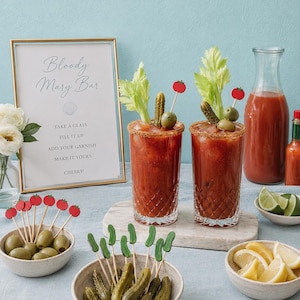 May include: Two tall crystal glasses filled with red Bloody Mary cocktails, garnished with celery, pickles, and tomato-topped picks. A framed sign reads Bloody Mary Bar, accompanied by bowls of extra pickles, olives, lemon wedges, and lime slices.