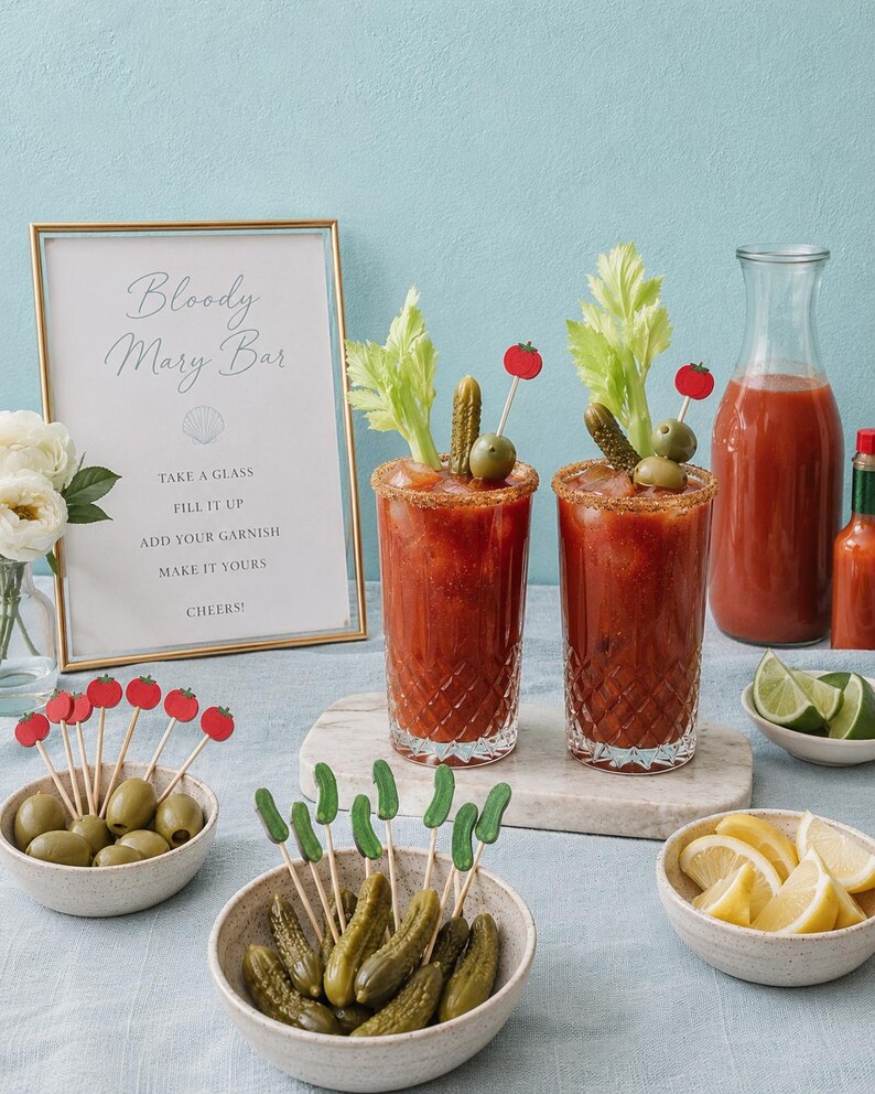 May include: Two tall crystal glasses filled with red Bloody Mary cocktails, garnished with celery, pickles, and tomato-topped picks. A framed sign reads Bloody Mary Bar, accompanied by bowls of extra pickles, olives, lemon wedges, and lime slices.