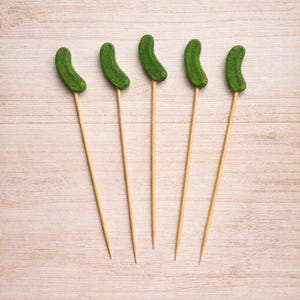 May include: Five wooden skewers, each topped with a small, textured, curved green pickle-shaped ornament. The skewers are thin, light-colored, and pointed at the bottom, while the green pickle toppers have a bumpy, realistic surface appearance.