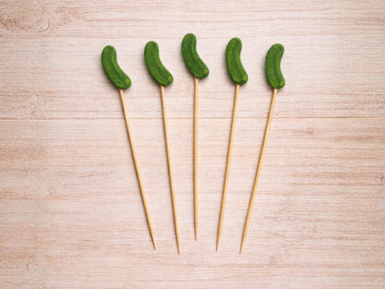 May include: Five wooden skewers, each topped with a small, textured, curved green pickle-shaped ornament. The skewers are thin, light-colored, and pointed at the bottom, while the green pickle toppers have a bumpy, realistic surface appearance.
