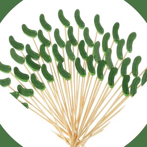 May include: A collection of wooden food picks, each topped with a small, curved, dark green pickle-shaped decoration. The light-colored wooden skewers are arranged in a fan shape, with the green pickle toppers pointing outward in various directions.