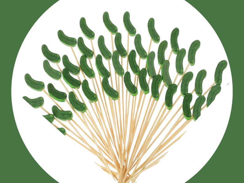 May include: A collection of wooden food picks, each topped with a small, curved, dark green pickle-shaped decoration. The light-colored wooden skewers are arranged in a fan shape, with the green pickle toppers pointing outward in various directions.