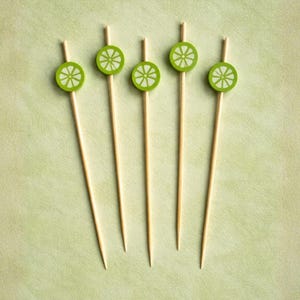 May include: Five wooden cocktail picks arranged vertically. Each pick features a small, circular lime slice topper in bright green with white segment details. The picks are thin, pointed, and light brown in colour, designed for garnishing drinks.