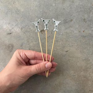 May include: Three cocktail picks with martini glass designs. Each pick features a white martini glass with black detailing and a green olive. The picks are held together and are made of wood.