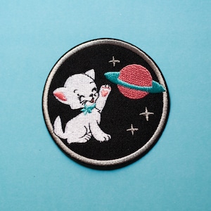 Może przedstawiać: Biały kot z różowym nosem i niebieskimi oczami sięga po różową planetę z turkusowym pierścieniem w czarnej przestrzeni z białymi gwiazdami. Obrazek jest okrągłą haftowaną naszywką.