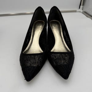 K&ouml;nnte beinhalten: Ein Paar schwarze Pumps mit Spitze und spitzer Zehenpartie. Die Innenseite der Schuhe ist goldfarben, die Abs&auml;tze sind niedrig. Die Schuhe stehen vor einem wei&szlig;en Hintergrund.
