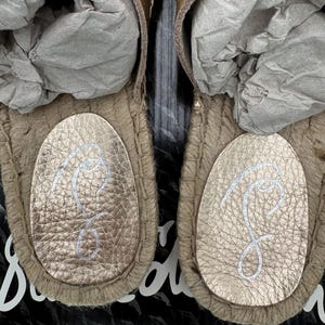May include: Close-up of two gold-colored sandals with a textured leather footbed and a woven jute sole. The footbed features a white cursive "S" logo. The sandals are new and stuffed with gray paper.