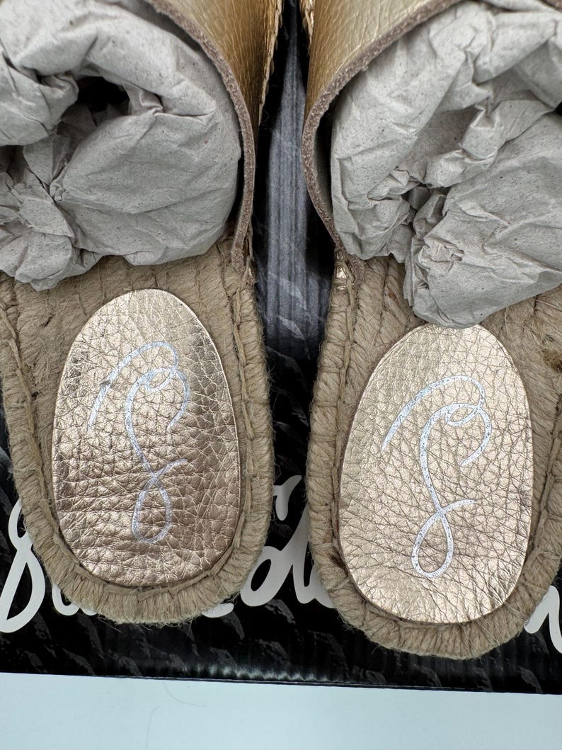 May include: Close-up of two gold-colored sandals with a textured leather footbed and a woven jute sole. The footbed features a white cursive "S" logo. The sandals are new and stuffed with gray paper.