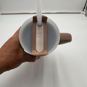 Pu&ograve; includere: Un bicchiere termico beige e bianco con una cannuccia trasparente. Il bicchiere ha un manico e un coperchio con un portacannuccia. Il coperchio e il manico sono di un colore beige pi&ugrave; scuro. Il bicchiere &egrave; progettato per bevande fredde.