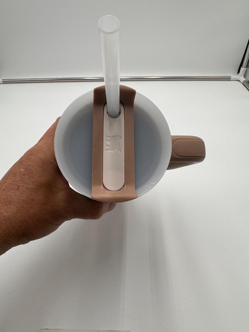 Pu&ograve; includere: Un bicchiere termico beige e bianco con una cannuccia trasparente. Il bicchiere ha un manico e un coperchio con un portacannuccia. Il coperchio e il manico sono di un colore beige pi&ugrave; scuro. Il bicchiere &egrave; progettato per bevande fredde.