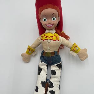 May include: A Jessie doll from the Toy Story franchise. The doll has a red hat, red hair, and a smiling face. She wears a cream-colored shirt with a yellow yoke, blue jeans, and cow-print pants. The doll's arms are outstretched.