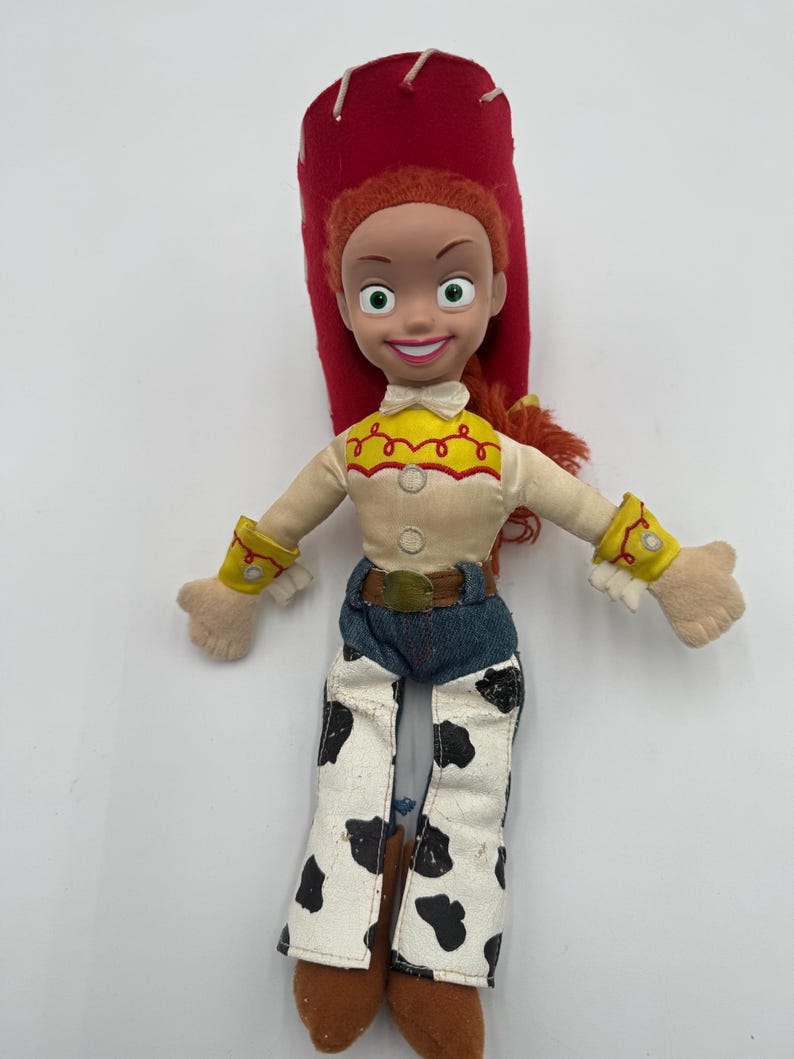 May include: A Jessie doll from the Toy Story franchise. The doll has a red hat, red hair, and a smiling face. She wears a cream-colored shirt with a yellow yoke, blue jeans, and cow-print pants. The doll's arms are outstretched.