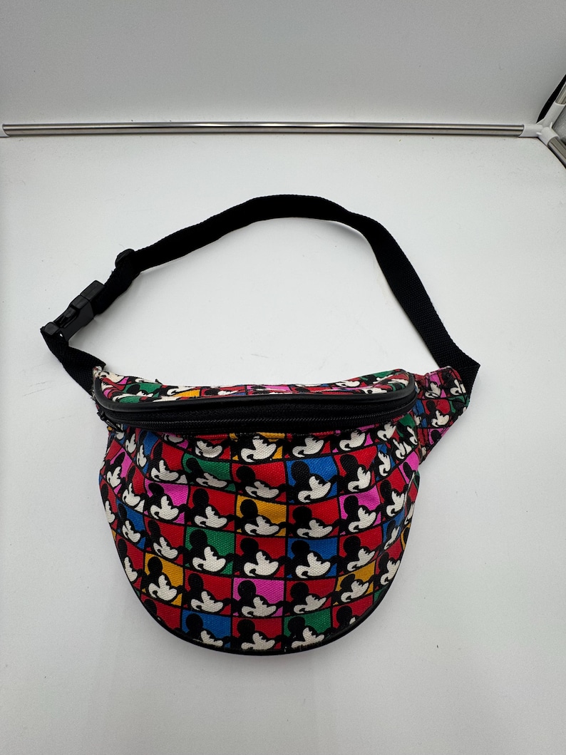 May include: A fanny pack featuring a colorful grid pattern of Mickey Mouse silhouettes in red, yellow, blue, green, and pink squares. The bag has a black zipper closure along the top and an adjustable black nylon strap with a plastic buckle.